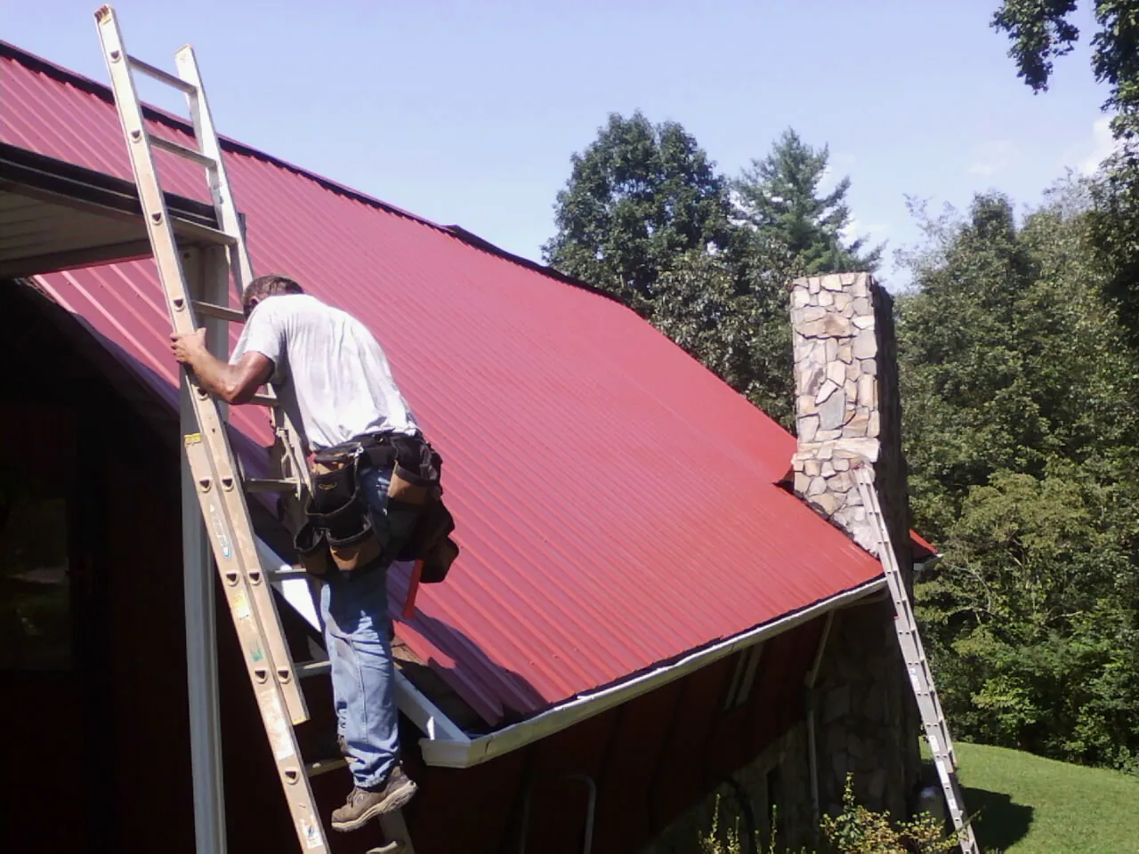 Roofer on a ladder beside a red metal roof and stone chimney — Soffit & Fascia Repair by Roofing Brothers in New Castle